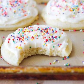 Soft sugar cookies frosted and lined up on a sheet pan with rainbow sprinkles. One has a bite taken out of it