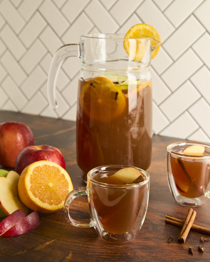 2 glasses of apple cider in front of a pitcher of cider with sliced apples and an orange