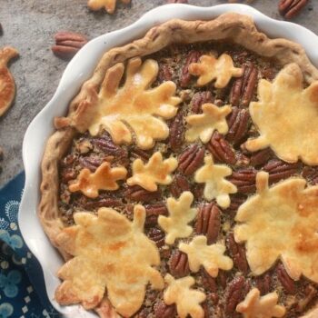 Salted maple pecan pie with decorative leaf pie pieces on top. Pie pieces and pecan are also laying around the pie.
