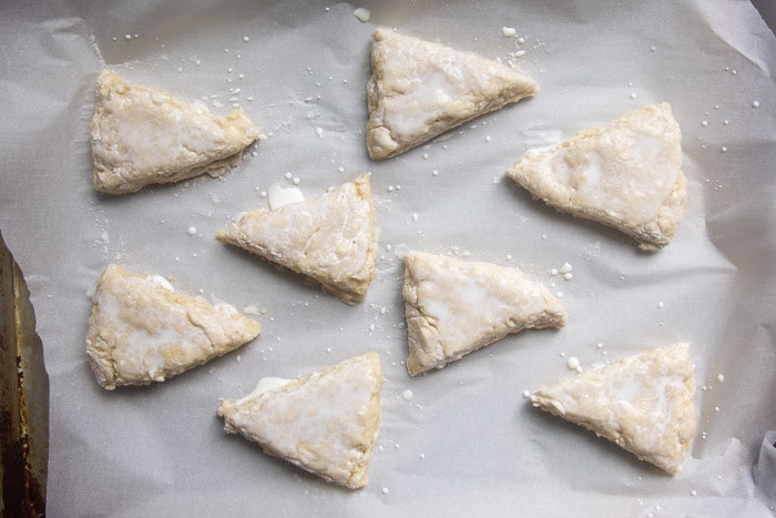 Learn the process of how to make lemon scones that are soft, fluffy, and moist! These lemon scones are tender and flavorful and topped with a tangy lemon glaze! Raw scone triangle brushed with cream about to be baked