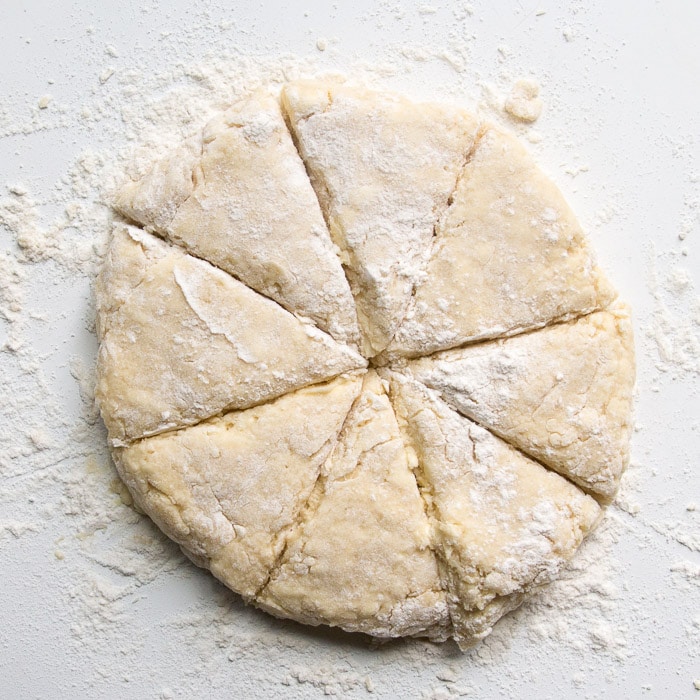 Learn the process of how to make lemon scones that are soft, fluffy, and moist! These lemon scones are tender and flavorful and topped with a tangy lemon glaze! Scone dough in a round shape cut into 8 trianges