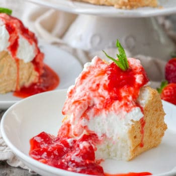 A piece of angel food cake topped with fresh strawberry sauce and a mint sprig, sitting in front of the cut-into angel food cake on a cake stand