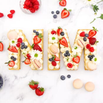 A beautiful cream tart in the shapes of an "N" and "M" decorated with flowers, fresh fruits and macarons