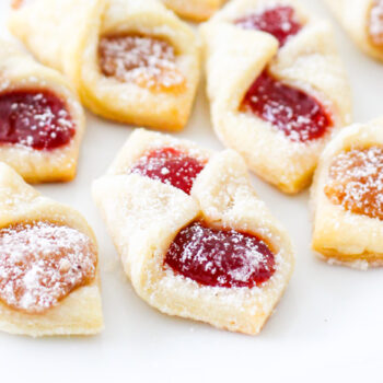 Showing the finished kolaczki cookies. They are on a white tray and you can see the cherry filling and almond filling in the cookies