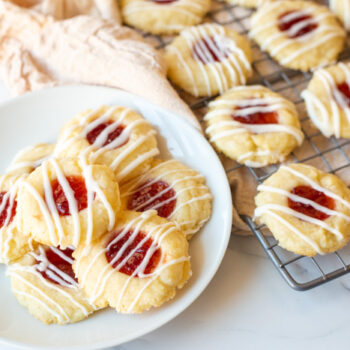Plate of finished shortbread thumbprint cookies with a cooling rack in the background with cookies on top