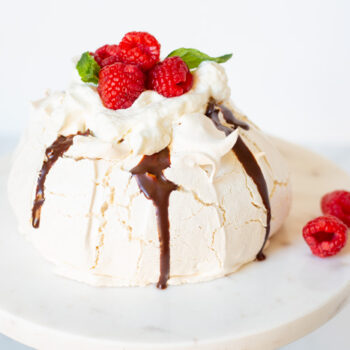 Pavlova topped with chocolate ganache, cream and fresh raspberries on a cake stand