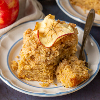 Slice of apple crumb cake on a plate with a bite on a fork and an apple chip on top