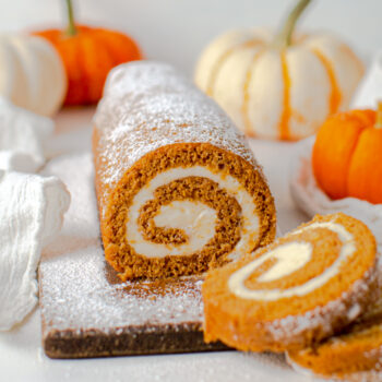 cream cheese pumpkin roll on a cutting board with 2 slices cut out and pumpkins in the background