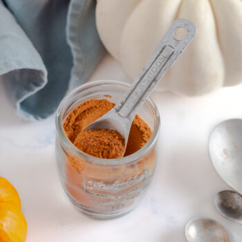 Jar of homemade pumpkin pie spice with a measuring spoon scooping some out and pumpkins in the background