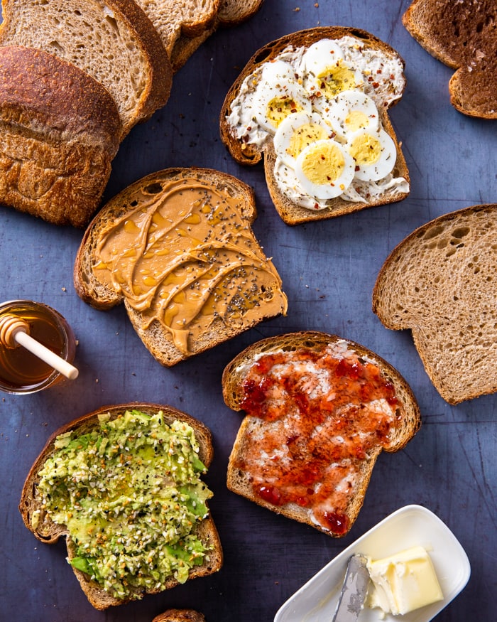 Slices of honey wheat sourdough topped with a variety of toppings including: avocado, cherry jam, peanut butter, and sliced hard boiled egg