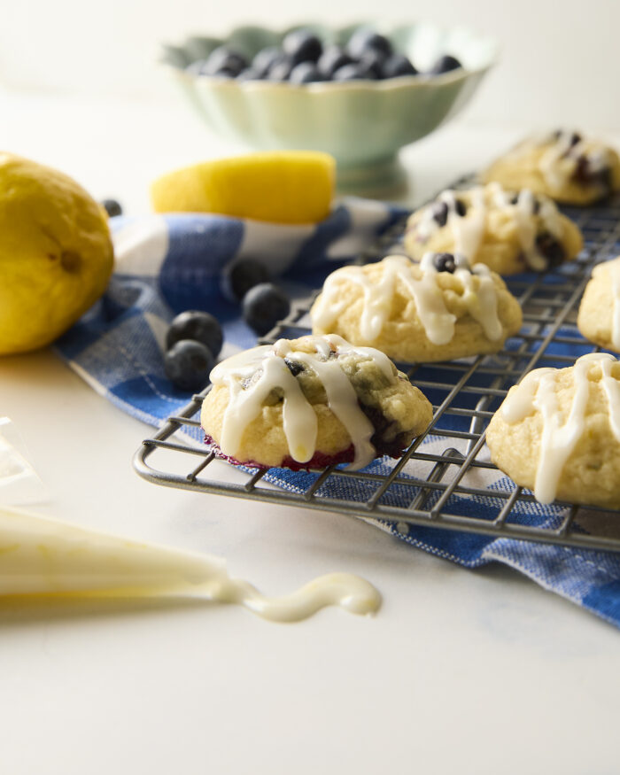 These blueberry cookies are soft, fluffy, and tender and are topped with a tangy lemon glaze! Perfect for summer gatherings! blueberry cookies with lemon glaze on a wire rack